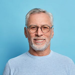 Portrait of handsome bearded European man with grey hair and beard smiles pleasantly looks directly at camera being in good mood has lucky day wears spectacles and sweater isolated over blue wall